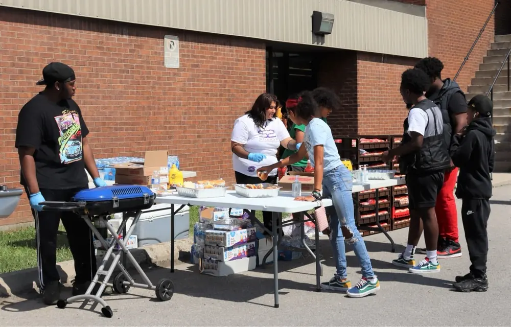Volunteers serving food to youth at an outdoor barbecue beside a grill